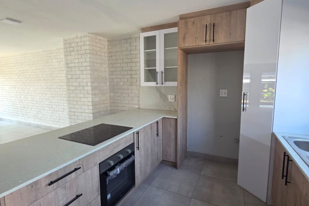Modern kitchen featuring light wood-look cabinetry, stone countertops, built-in oven, electric hob, and large format floor tiles, adjacent to an exposed brick living area.