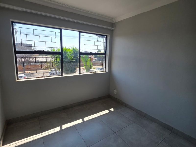 A bright, empty bedroom featuring neutral grey tiled flooring, clean walls, and large windows fitted with security bars.