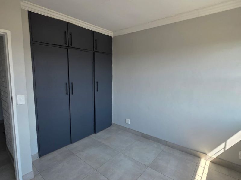 A bright, empty bedroom featuring modern grey built-in cupboards, neutral tiled flooring, and clean finishes throughout.