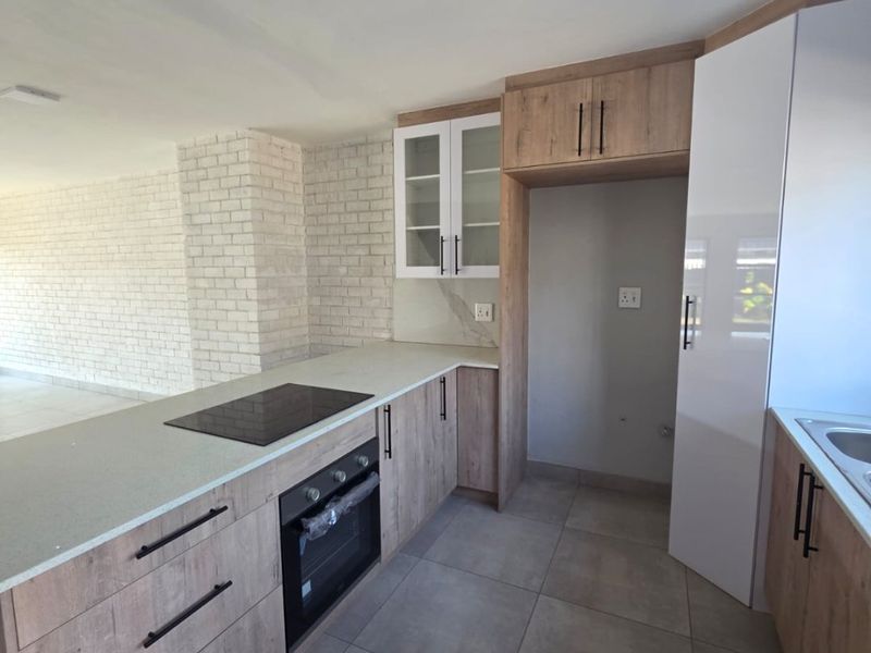 Modern kitchen featuring light wood-look cabinetry, stone countertops, built-in oven, electric hob, and large format floor tiles, adjacent to an exposed brick living area.