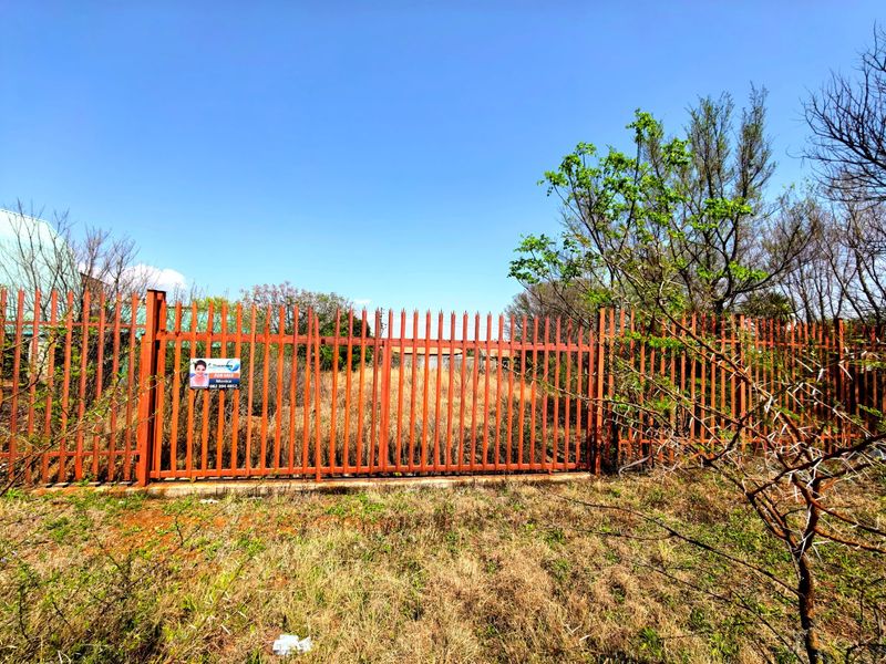 A vacant residential plot secured by steel palisade fencing and a gate, featuring overgrown vegetation and clear street-level access.