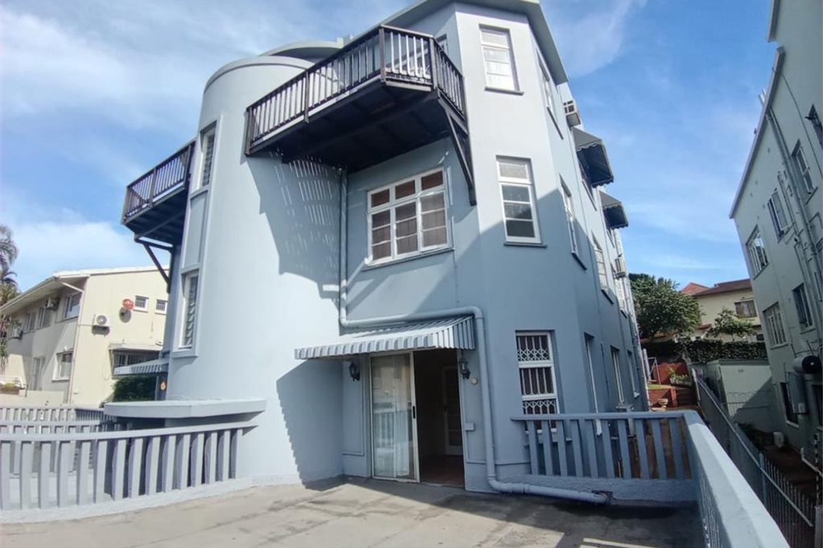 Rear exterior view of the apartment building featuring multiple balconies, sliding glass doors, and protective awnings with a paved outdoor area.