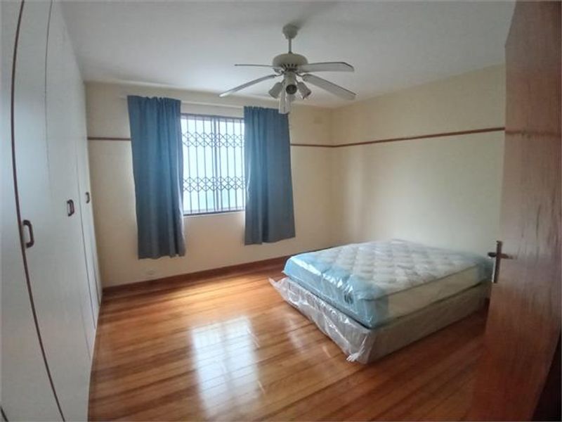 A spacious bedroom featuring polished hardwood flooring, built-in cupboards, a ceiling fan, and a window fitted with a security gate.