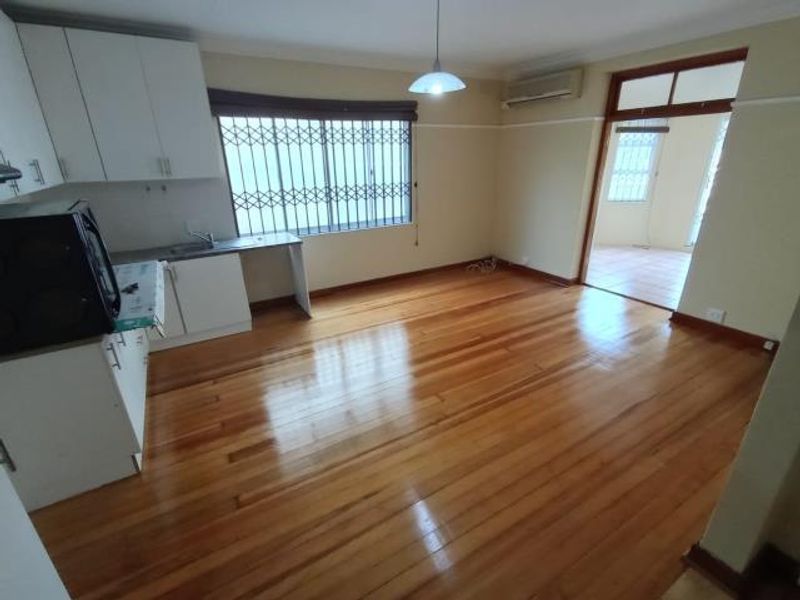 An open-plan living and kitchen area featuring polished hardwood floors, white cabinetry, and a split-system air conditioner, offering a bright and versatile living space.