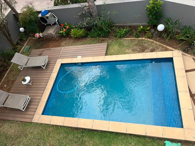An elevated view of a private swimming pool featuring tiled coping, adjacent timber decking, and a landscaped garden area with boundary wall.
