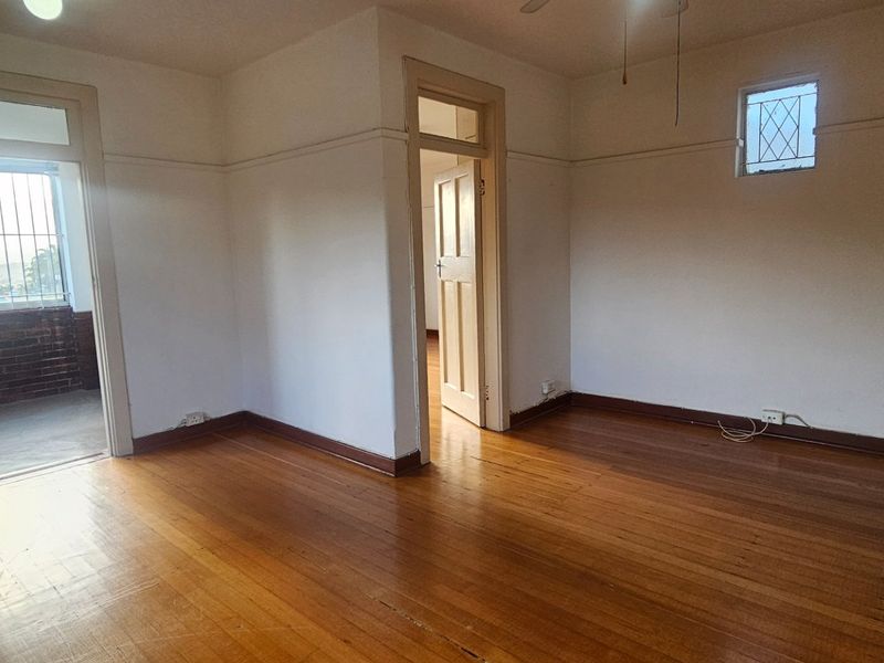 An empty living room featuring original hardwood flooring, timber door frames, and a small leaded glass window, offering a blank canvas for renovation.