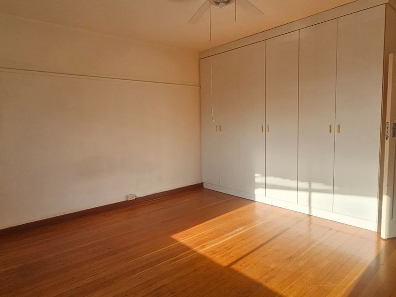 A bright, empty bedroom featuring polished hardwood flooring, extensive built-in cupboards, and a ceiling fan, offering a clean and neutral space.