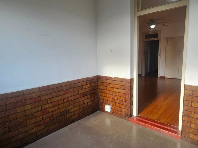 An enclosed patio area featuring brick wainscoting and timber flooring, leading into an interior living space with built-in cupboards and a ceiling fan.