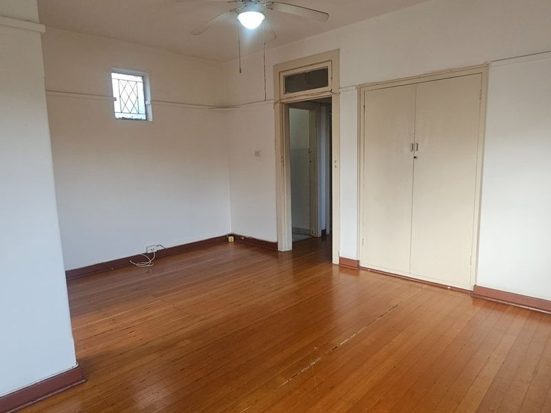 An empty bedroom featuring original timber flooring, a ceiling fan, and a built-in wardrobe, offering a blank canvas for renovation.