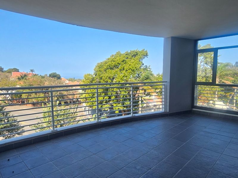 A spacious, covered balcony featuring dark tiled flooring, a modern metal balustrade, and expansive glass doors offering pleasant neighborhood and treetop views.