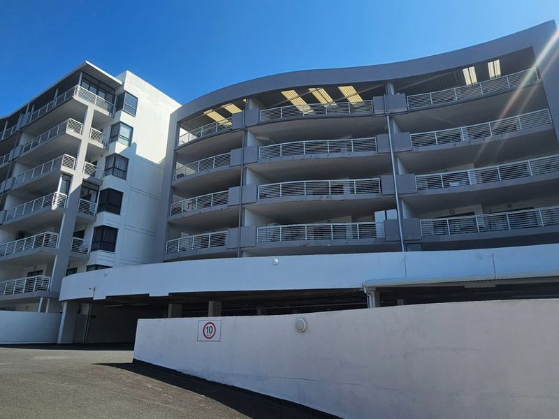 A modern multi-story apartment building featuring curved balconies with metal railings, a paved driveway, and a covered parking area.