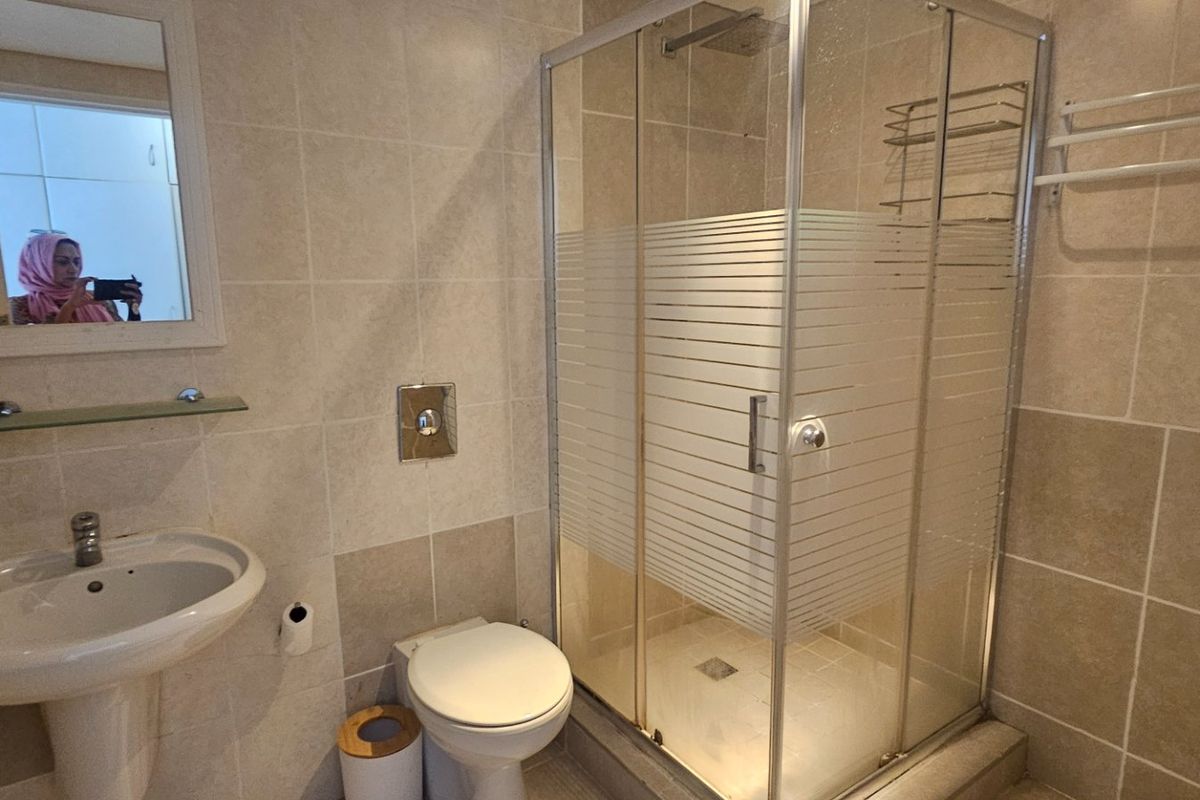 A functional bathroom featuring a pedestal basin, wall-mounted toilet, and a glass-enclosed shower with neutral wall and floor tiling.