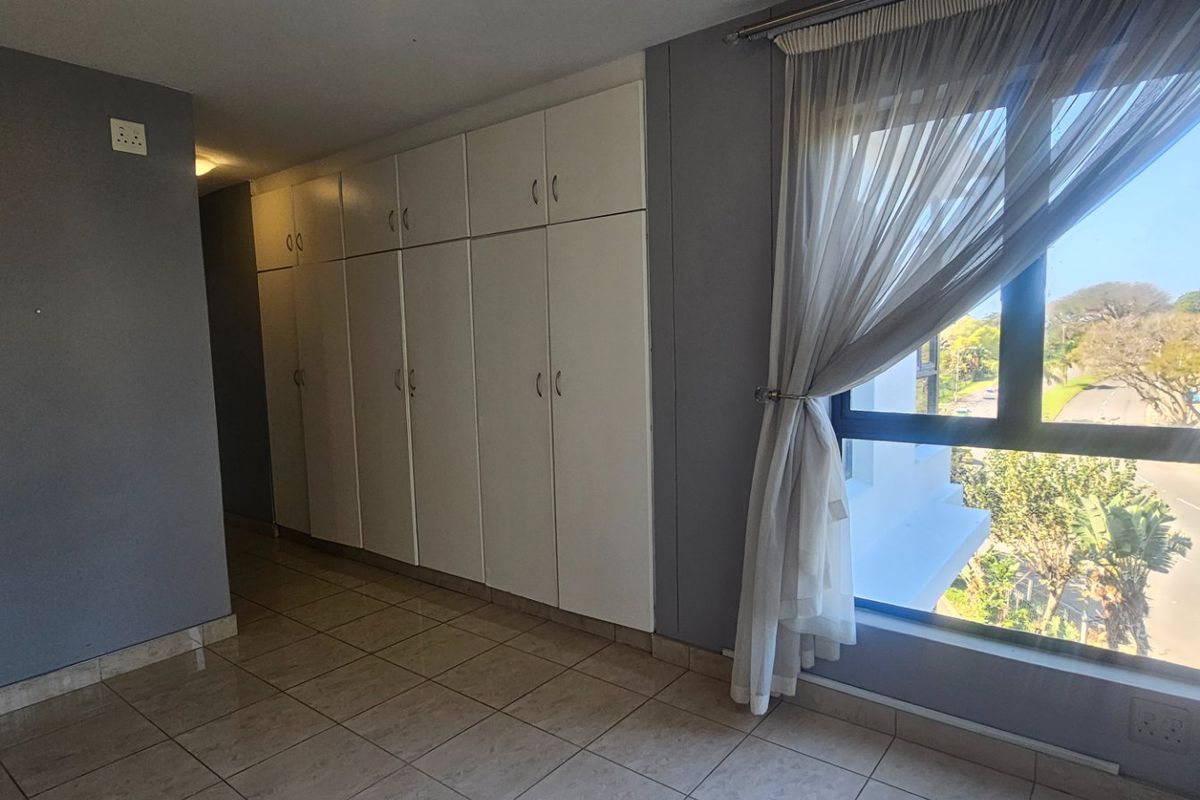 A bright, empty bedroom featuring tiled flooring, ample built-in cupboard storage, and a large window with natural light.