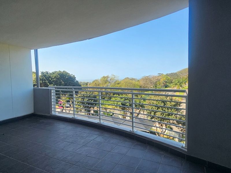 A spacious covered balcony featuring tiled flooring and a metal balustrade, offering scenic views of the surrounding greenery.