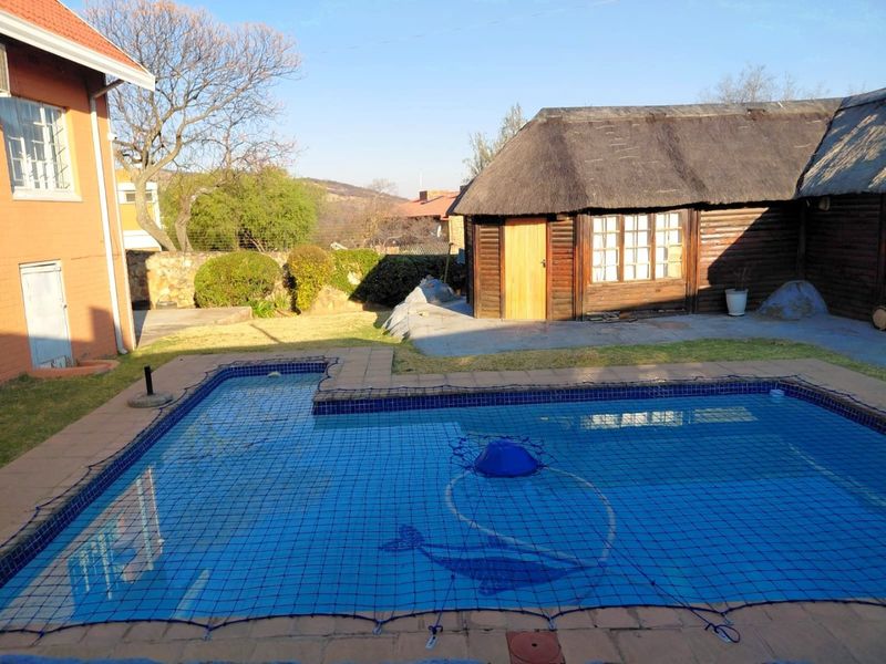 Outdoor pool area featuring a tiled swimming pool with safety net, adjacent to a rustic thatched-roof outbuilding and landscaped garden space.