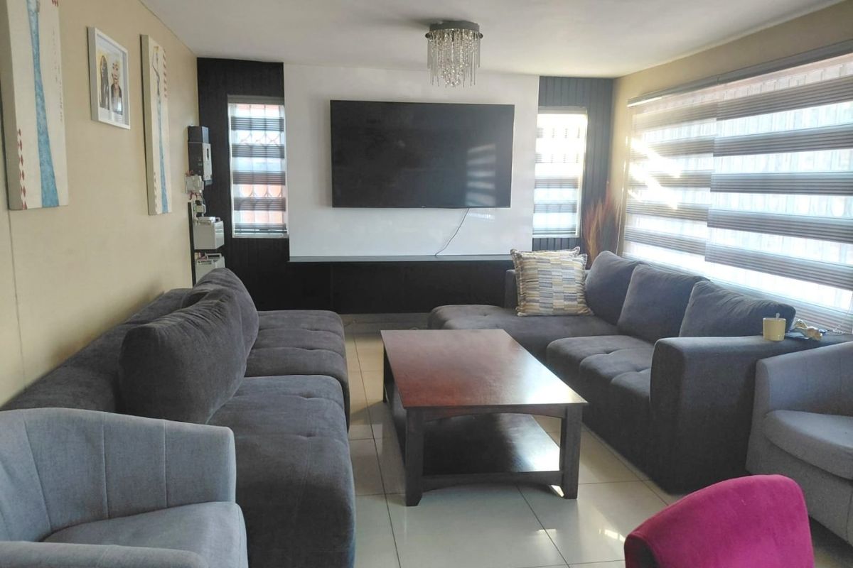 A comfortable living room featuring tiled flooring, a modern built-in media wall, and large windows with fitted roller blinds.