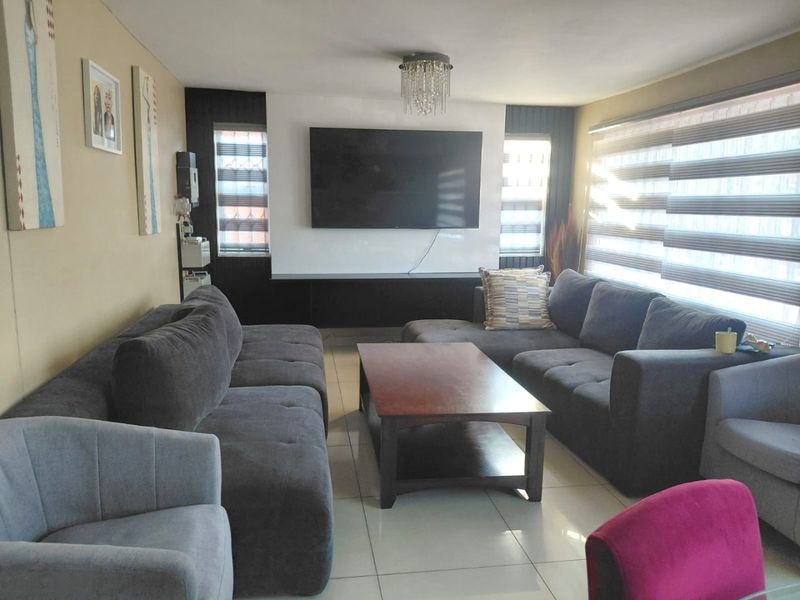 A comfortable living room featuring tiled flooring, a modern built-in media wall, and large windows with fitted roller blinds.
