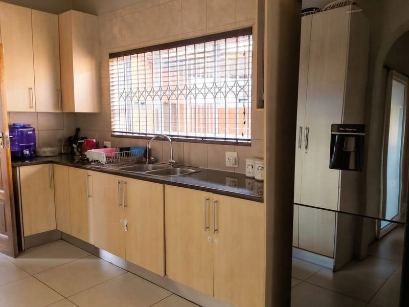 A functional kitchen featuring light wood-finish cabinetry, dark countertops, a double sink, and tiled flooring with natural light from a window with security bars.