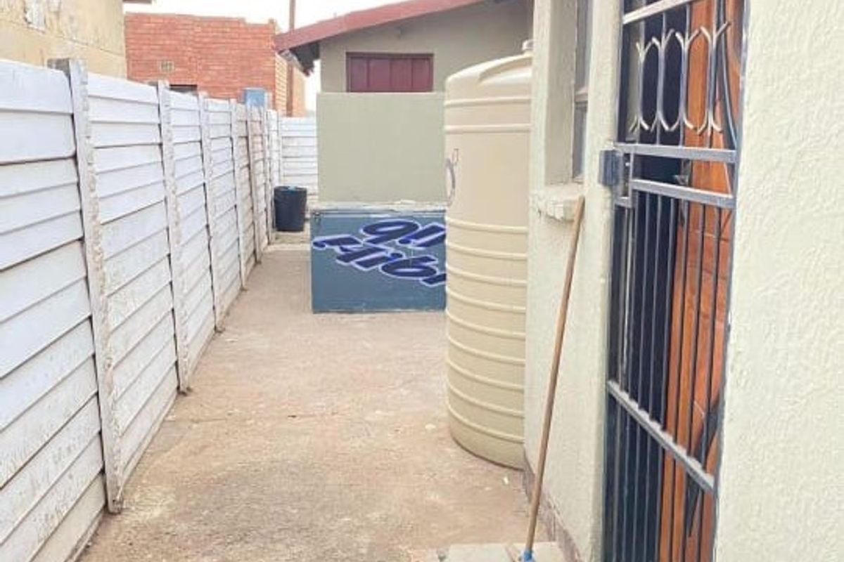A narrow side passage featuring a precast concrete boundary wall, a water storage tank, and a security gate entrance.