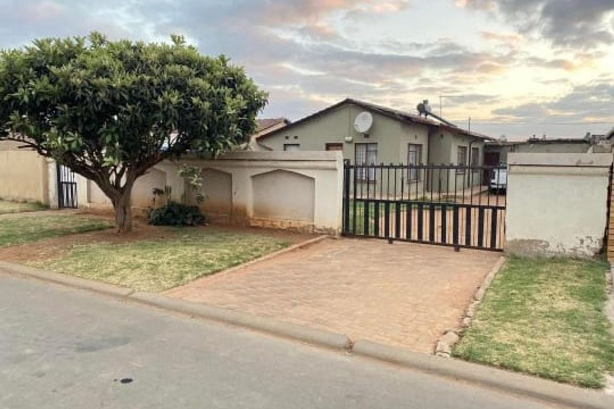 Front view of a residential house featuring a paved driveway, metal security gate, perimeter wall, and established garden area.
