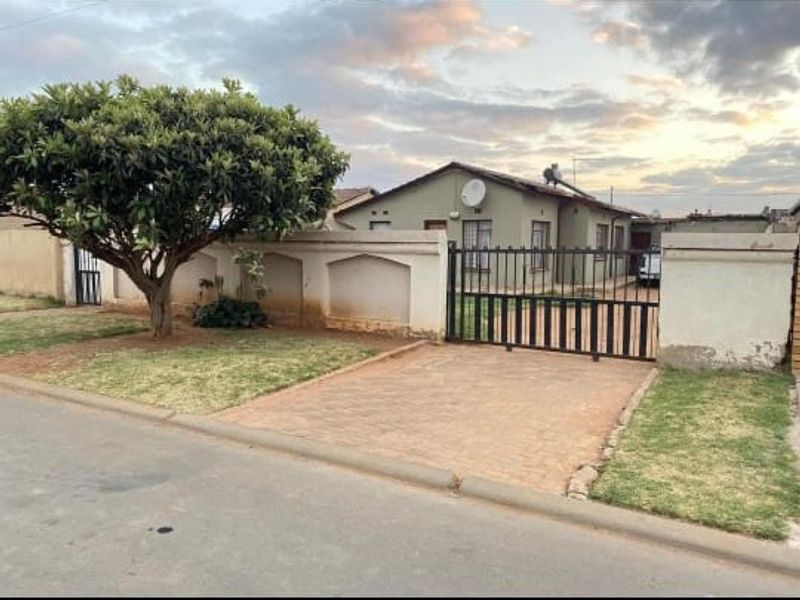 Front view of a residential house featuring a paved driveway, metal security gate, perimeter wall, and established garden area.