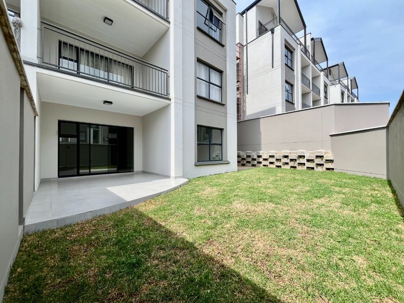 A modern apartment exterior featuring a private tiled patio, a small lawn area, and upper-level balconies with metal railings.
