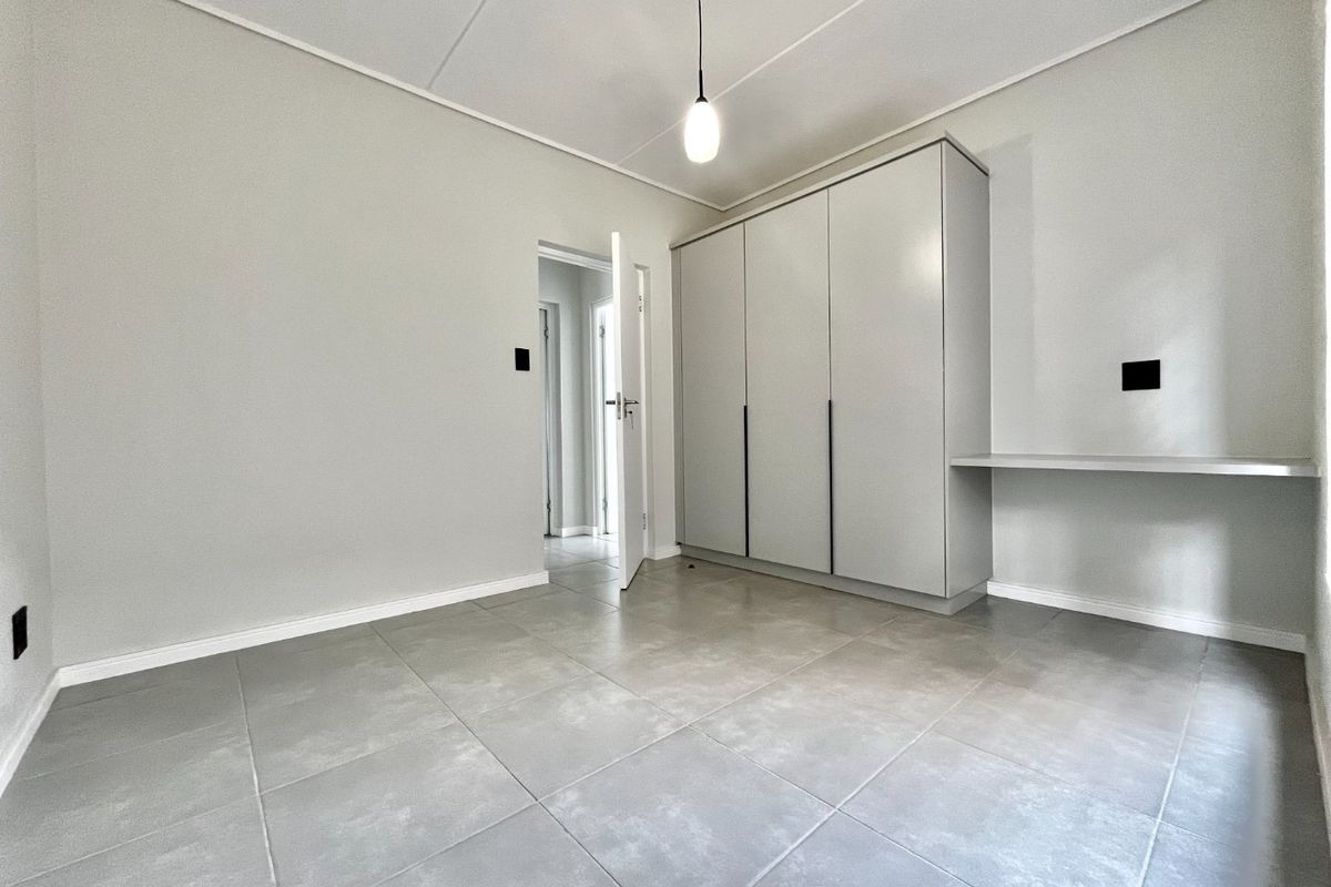 A clean, modern bedroom featuring neutral tiled flooring, a built-in wardrobe, and a functional built-in desk area with contemporary lighting.
