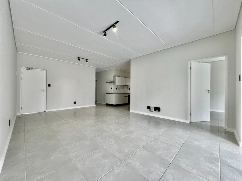 Spacious open-plan living area featuring neutral ceramic floor tiles, modern track lighting, and a clean, minimalist aesthetic leading into a functional kitchen space.