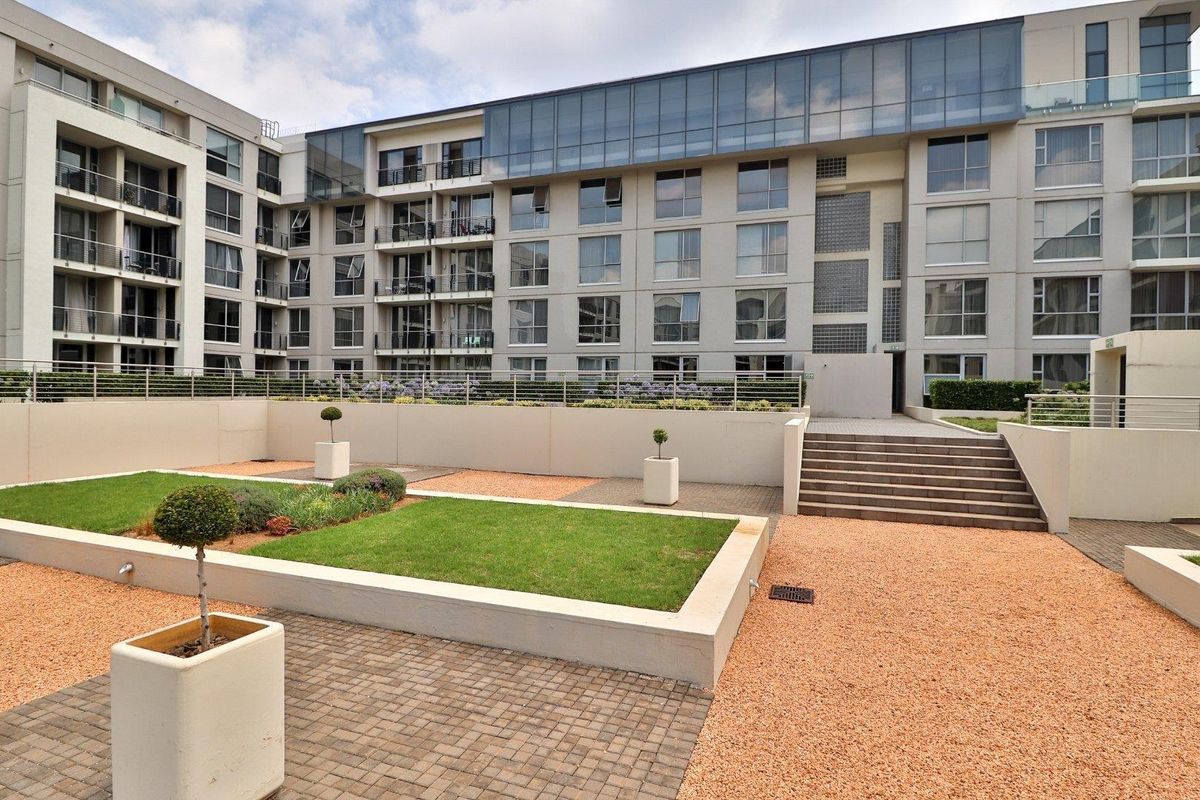 A well-maintained rear courtyard area featuring paved walkways, landscaped garden beds, and concrete stairs leading to a modern apartment building facade.