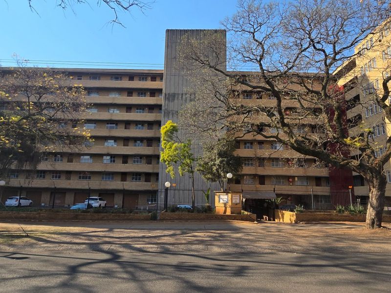 Street-level view of a multi-story apartment building featuring a brick facade, central concrete stairwell tower, and perimeter security fencing.