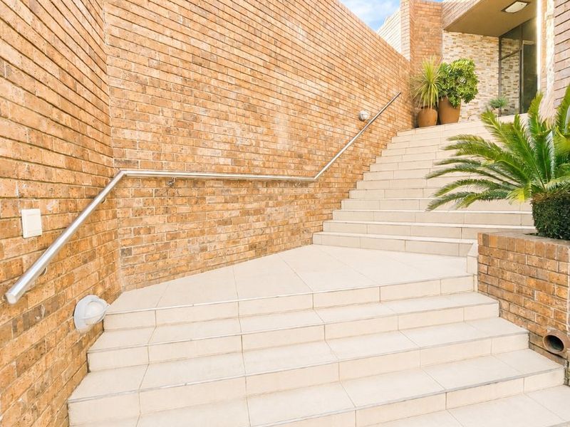 A well-maintained exterior entrance featuring a brick retaining wall, tiled staircase with a stainless steel handrail, and integrated wall lighting.