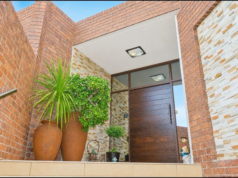 A well-maintained front entrance featuring a modern timber door, stone cladding accents, and a tiled landing with a stainless steel handrail.