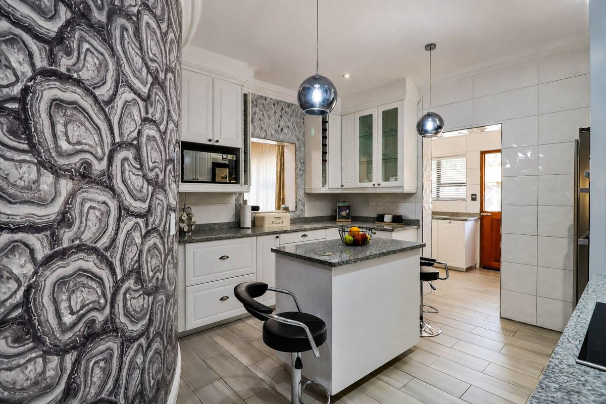 A well-appointed kitchen featuring white cabinetry, granite countertops, a central island, and modern pendant lighting, with an adjacent scullery area.