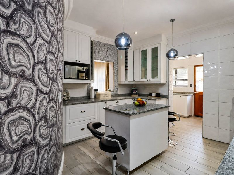 A well-appointed kitchen featuring white cabinetry, granite countertops, a central island, and modern pendant lighting, with an adjacent scullery area.