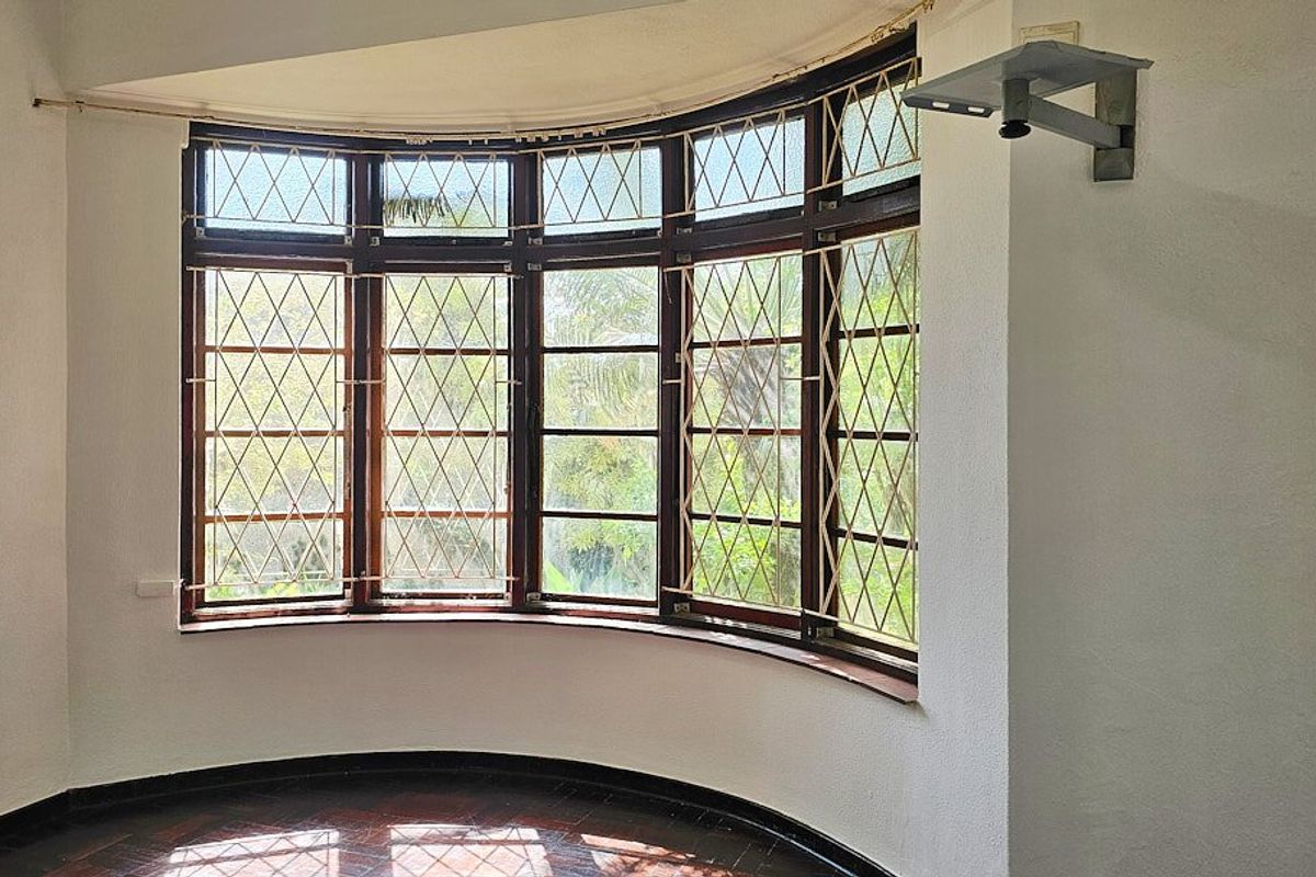 A bright, empty living room featuring a large curved bay window with timber frames, security bars, and original hardwood flooring.