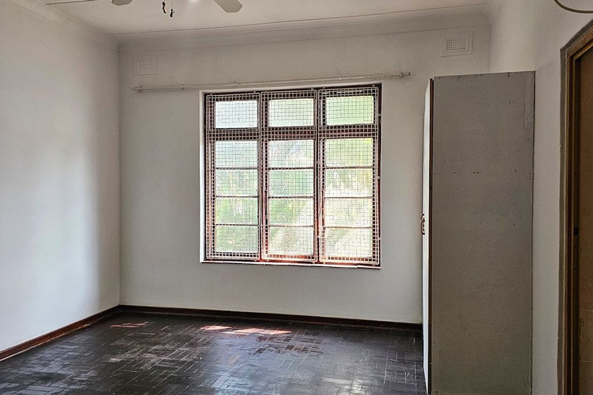 An empty bedroom featuring dark parquet flooring, a built-in wardrobe, and a large window fitted with security bars.