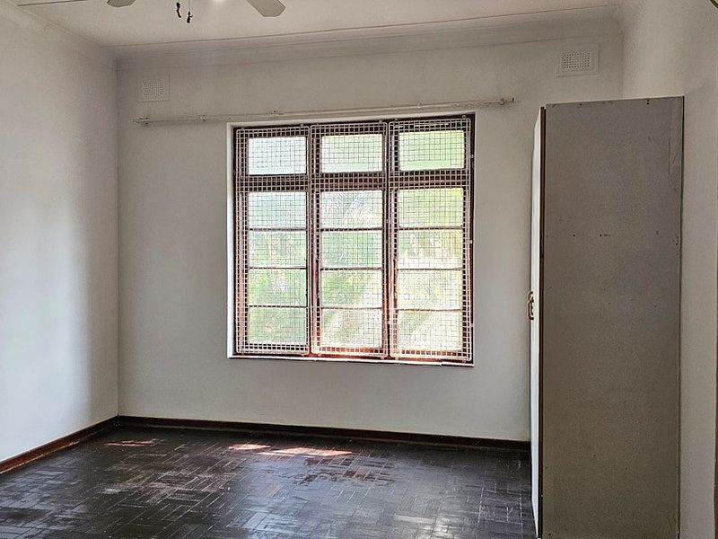 An empty bedroom featuring dark parquet flooring, a built-in wardrobe, and a large window fitted with security bars.