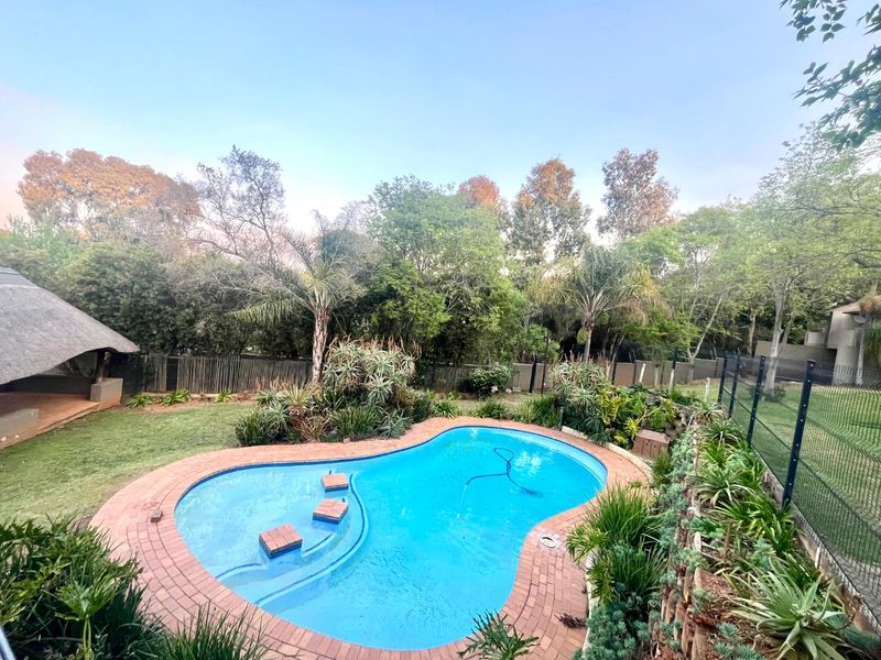 A well-maintained outdoor pool area featuring a kidney-shaped swimming pool with brick coping, surrounded by a lush garden and a thatched lapa.
