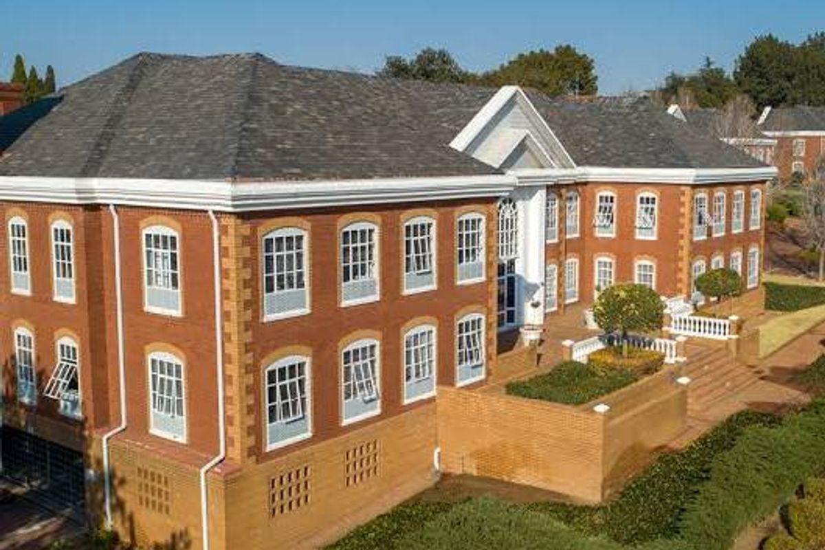 Aerial view of a multi-story commercial brick building featuring a pitched roof, extensive casement windows, and landscaped terraced gardens with retaining walls.