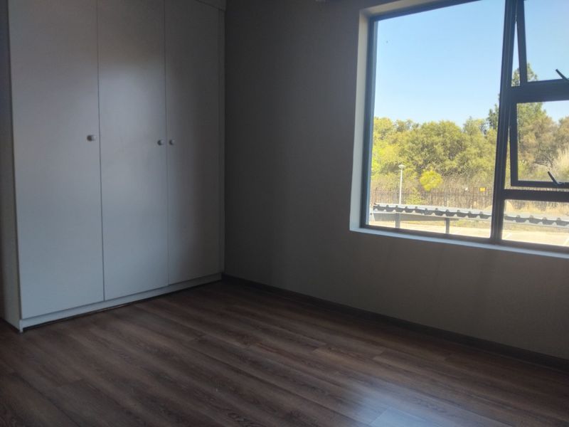 A bright, empty bedroom featuring modern laminate flooring, a built-in wardrobe, and a large window with natural light.