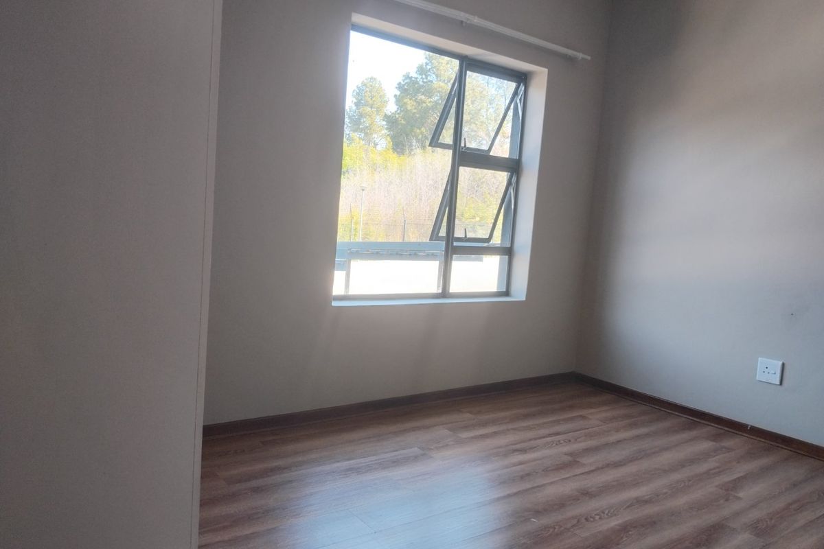 A bright, empty bedroom featuring modern laminate flooring and a large aluminium-framed window with natural light.