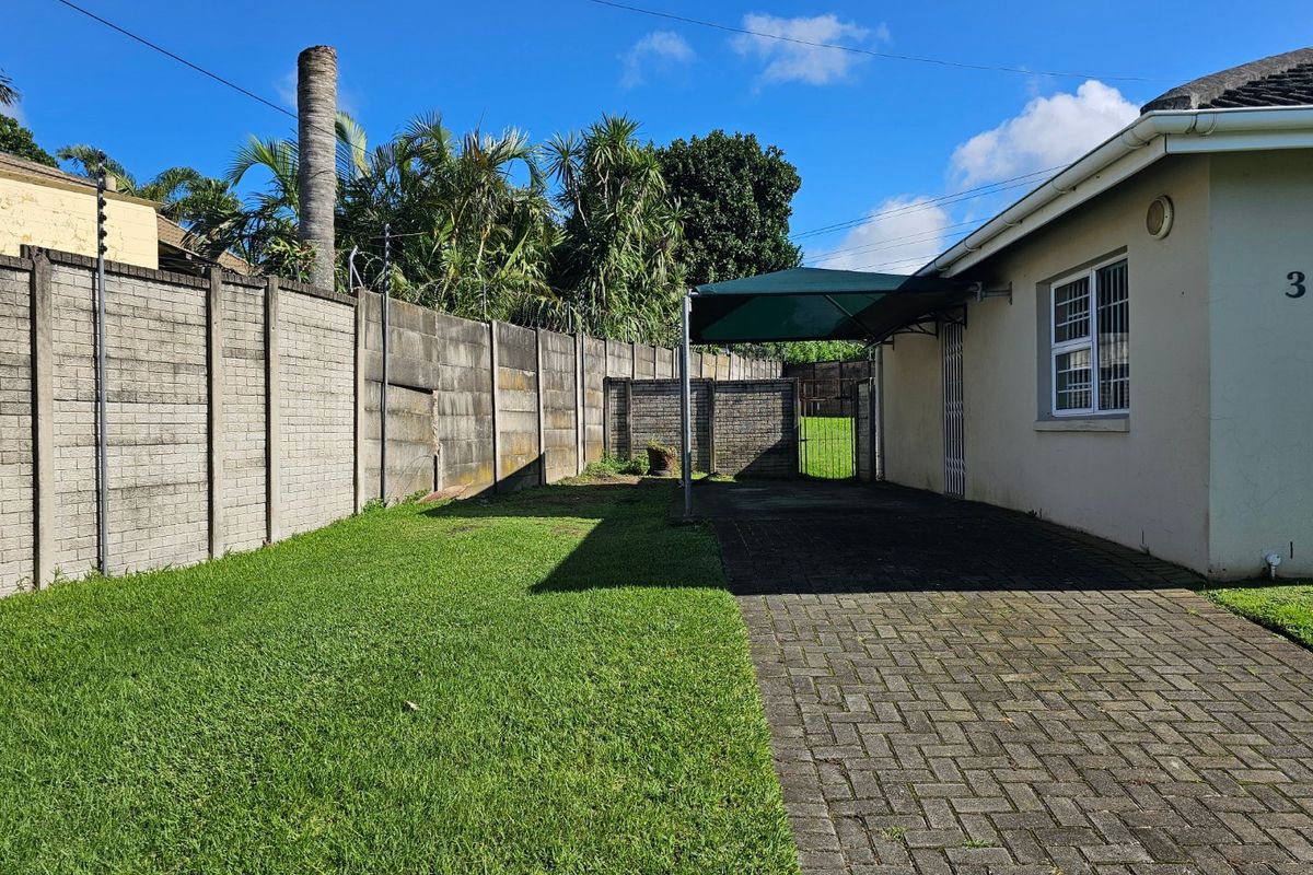 Rear exterior view of the townhouse featuring a paved driveway, carport, lawn area, and secure boundary wall with electric fencing.