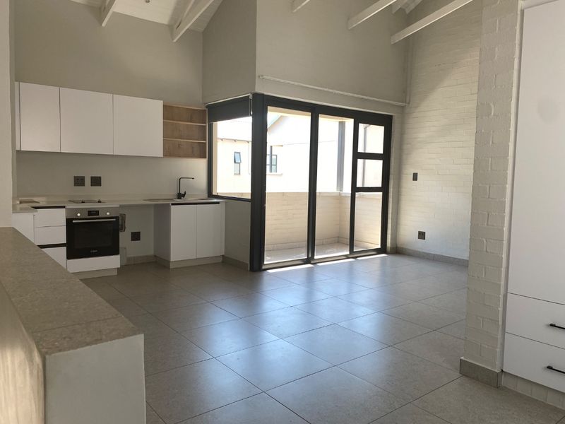 Modern open-plan living area featuring a sleek kitchen with built-in appliances, tiled floors, exposed rafters, and large sliding doors leading to a balcony.