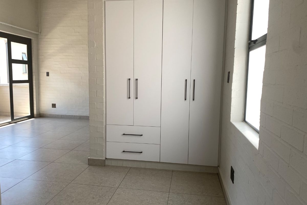 A bright, empty bedroom featuring modern built-in wardrobes, neutral tiled flooring, and painted brick walls with access to an outdoor area.