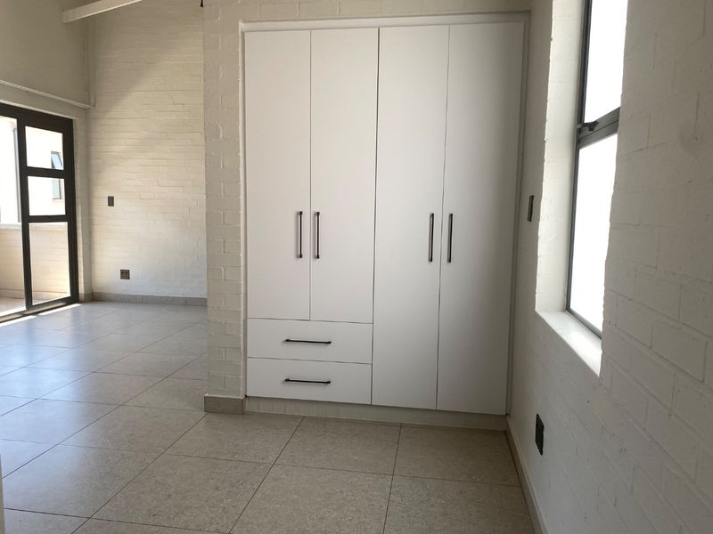 A bright, empty bedroom featuring modern built-in wardrobes, neutral tiled flooring, and painted brick walls with access to an outdoor area.