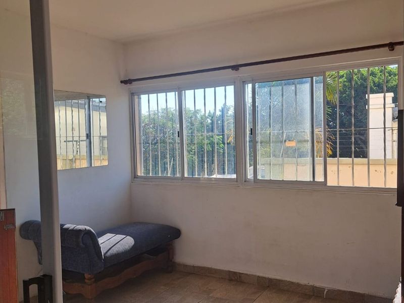 A sunlit enclosed balcony or patio area featuring tiled flooring, large windows with security bars, and a sliding glass door entrance.