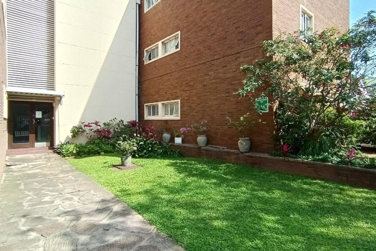 A well-maintained rear exterior view of the apartment building featuring a brick facade, stone paved walkway, and a landscaped garden area.