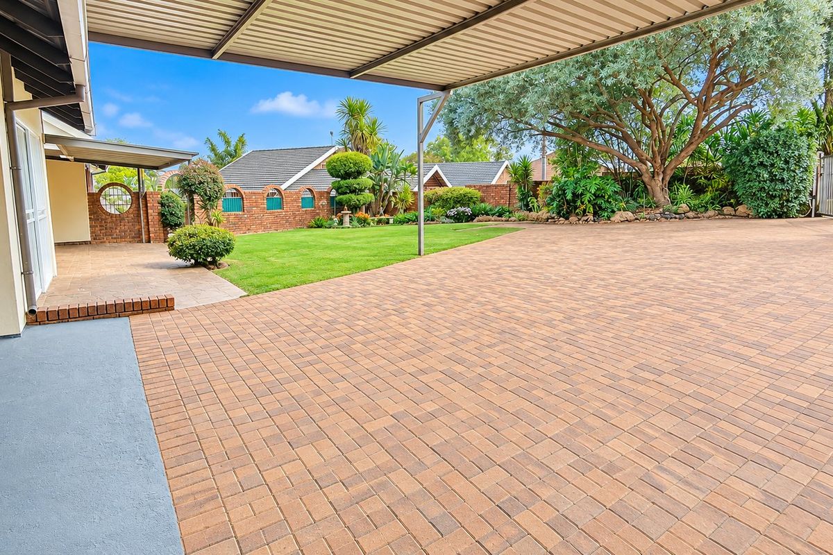 Spacious paved driveway and rear garden area featuring a covered patio, well-maintained lawn, and established landscaping with brick boundary walls.