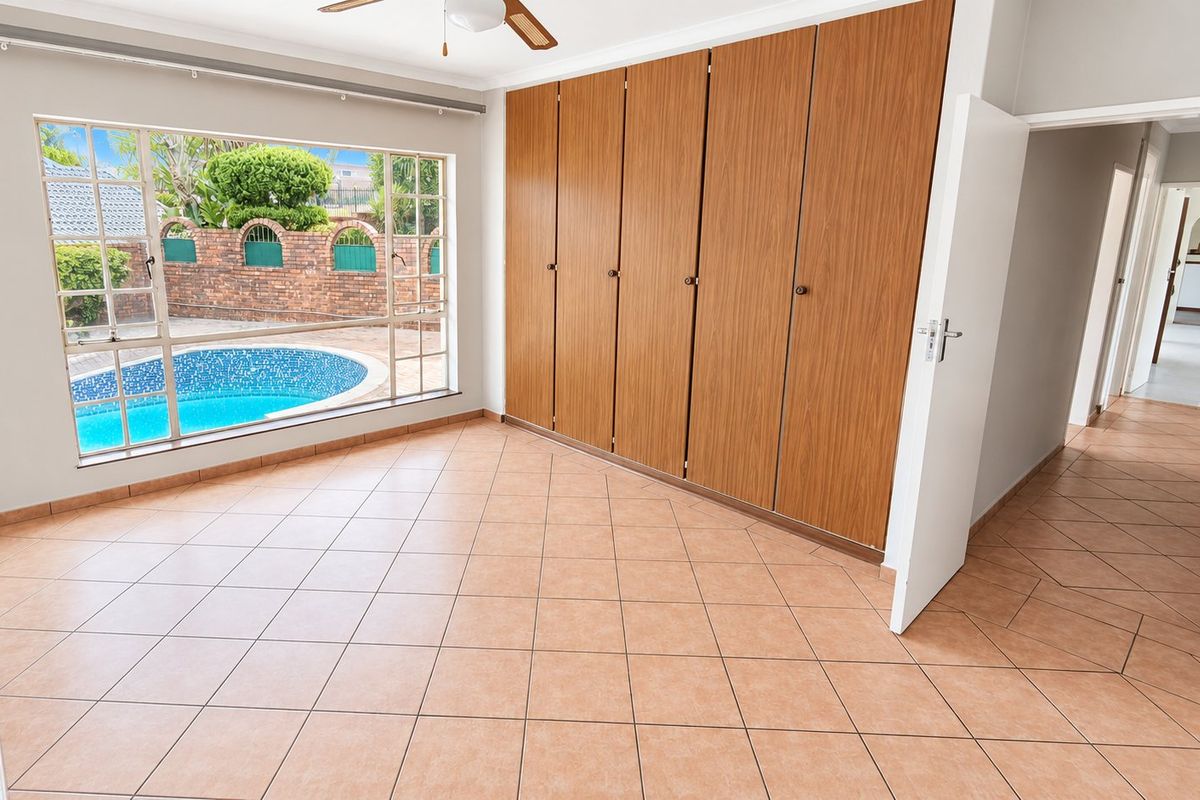 An empty bedroom featuring built-in wooden cupboards, tiled flooring, a ceiling fan, and a large window overlooking the swimming pool area.
