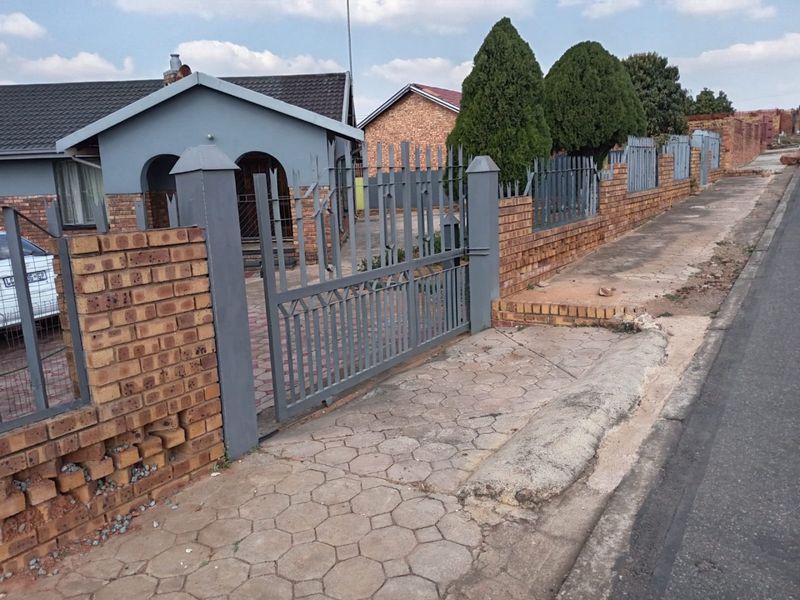 Street view of the property featuring a brick boundary wall, metal security gate, and paved driveway access from the sidewalk.